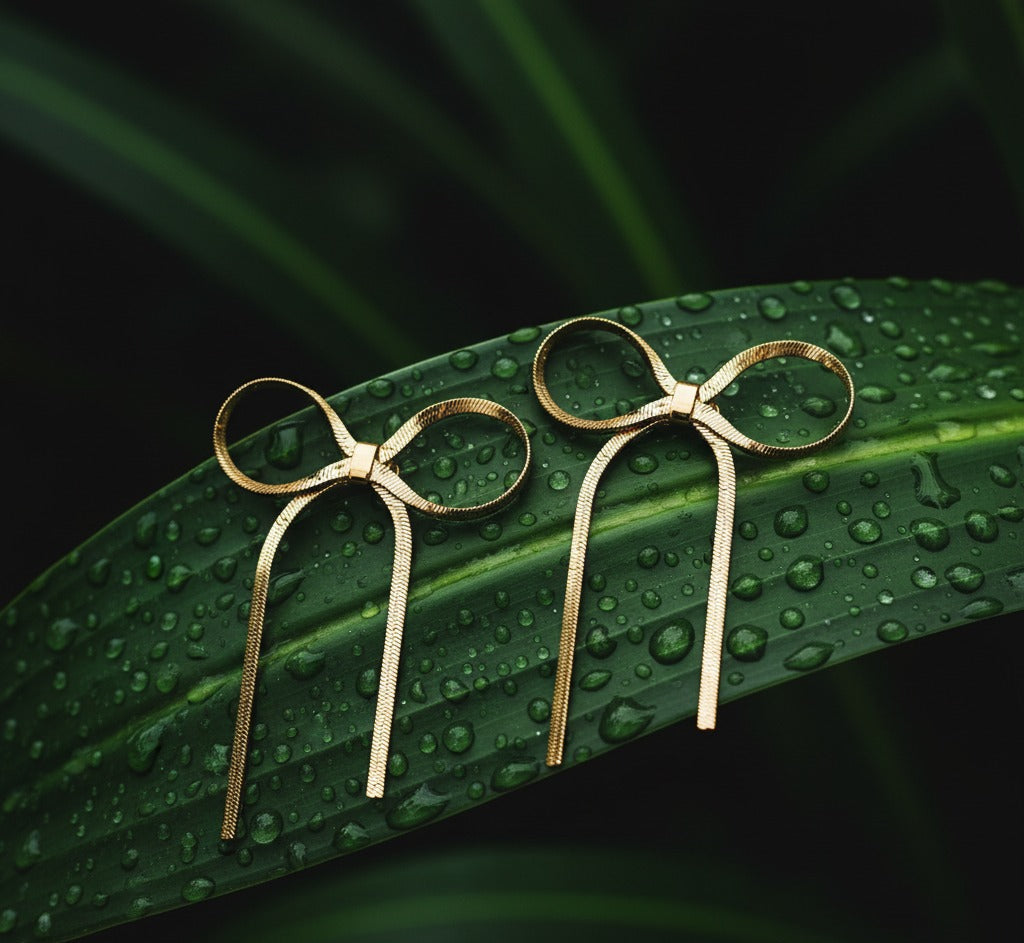 Gold bow-shaped earrings on a green leaf with water droplets