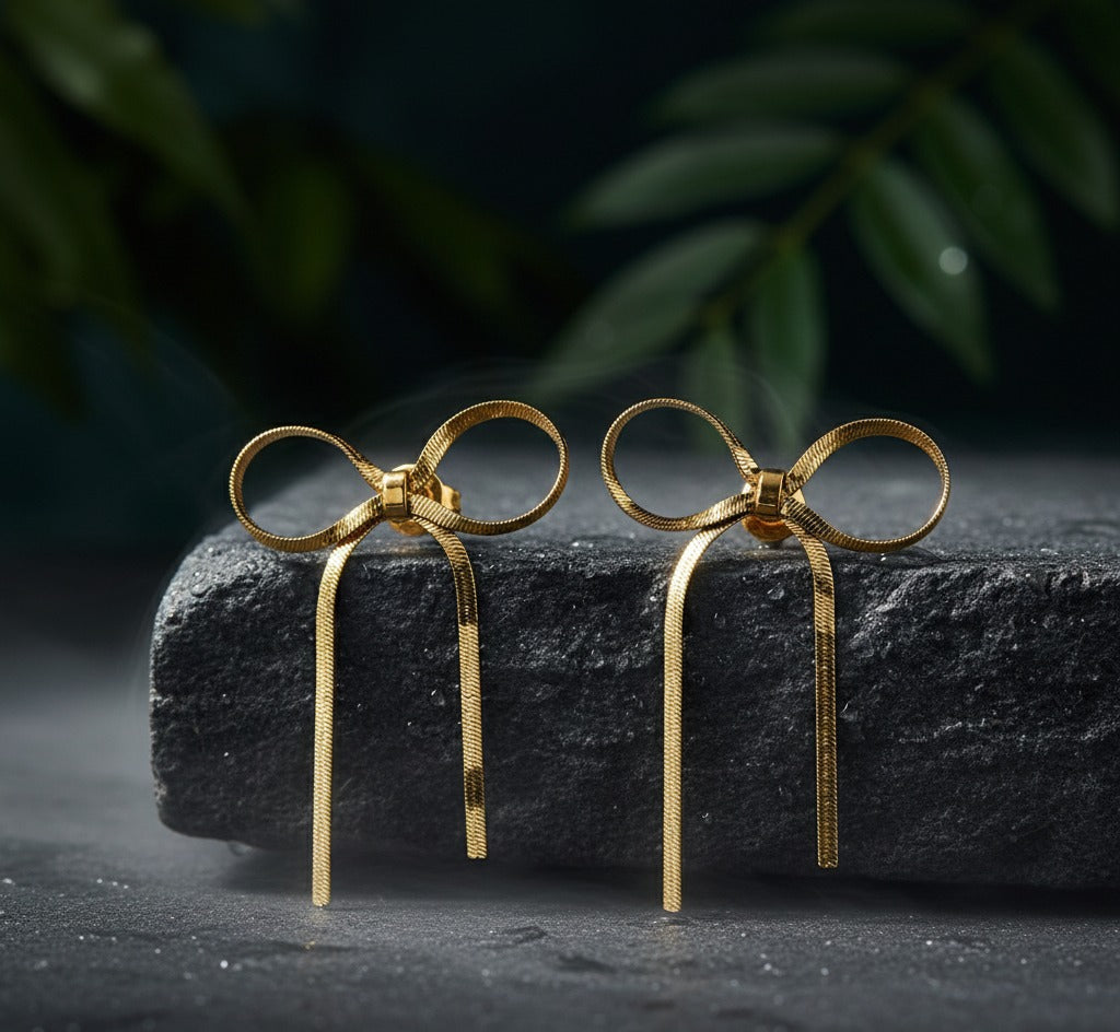 Gold bow-shaped earrings on a dark stone surface with a blurred green leaf background